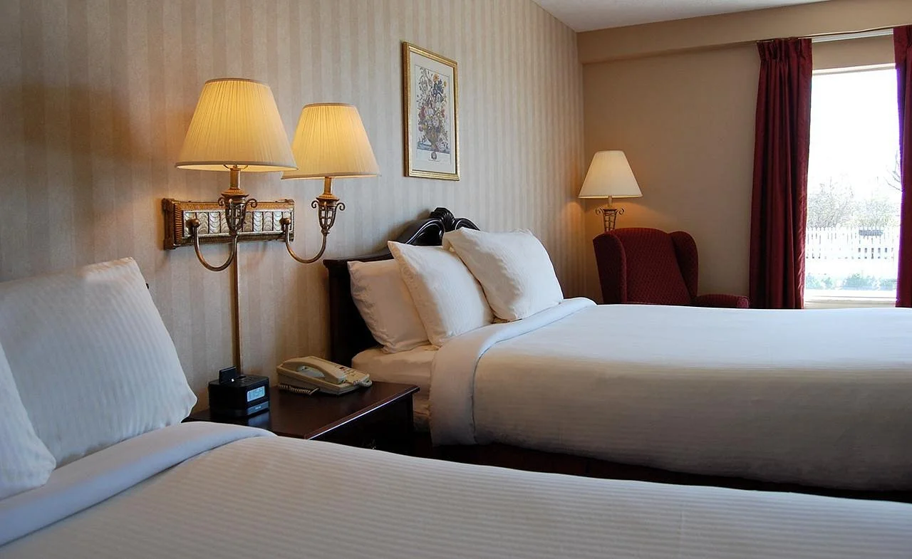Two double beds with white bedding, bedside lamp, and a window with red curtains in a hotel room.