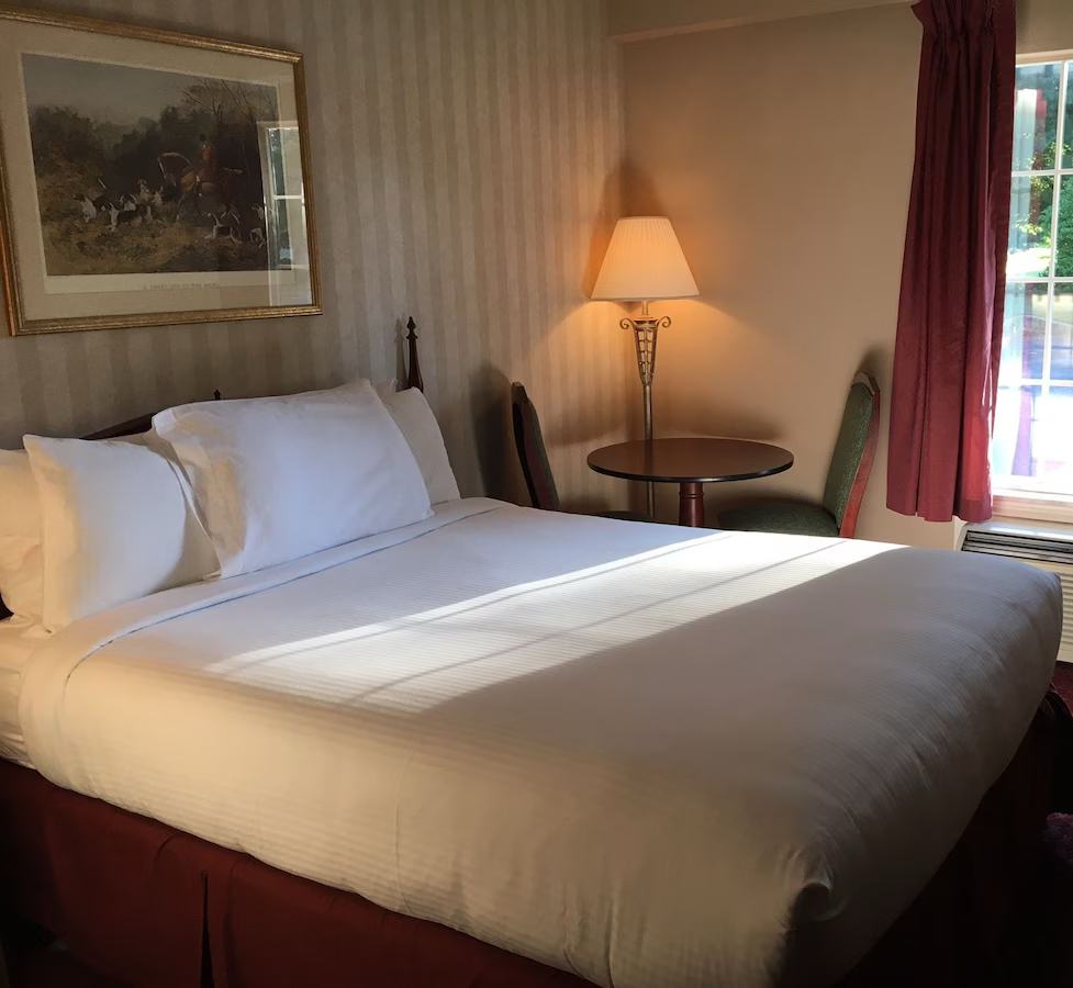 A neatly made bed with white bedding and pillows in a hotel room. There is a round wooden table with a lamp and two chairs beside a window with red curtains, and a painting hanging on the wall above the bed.