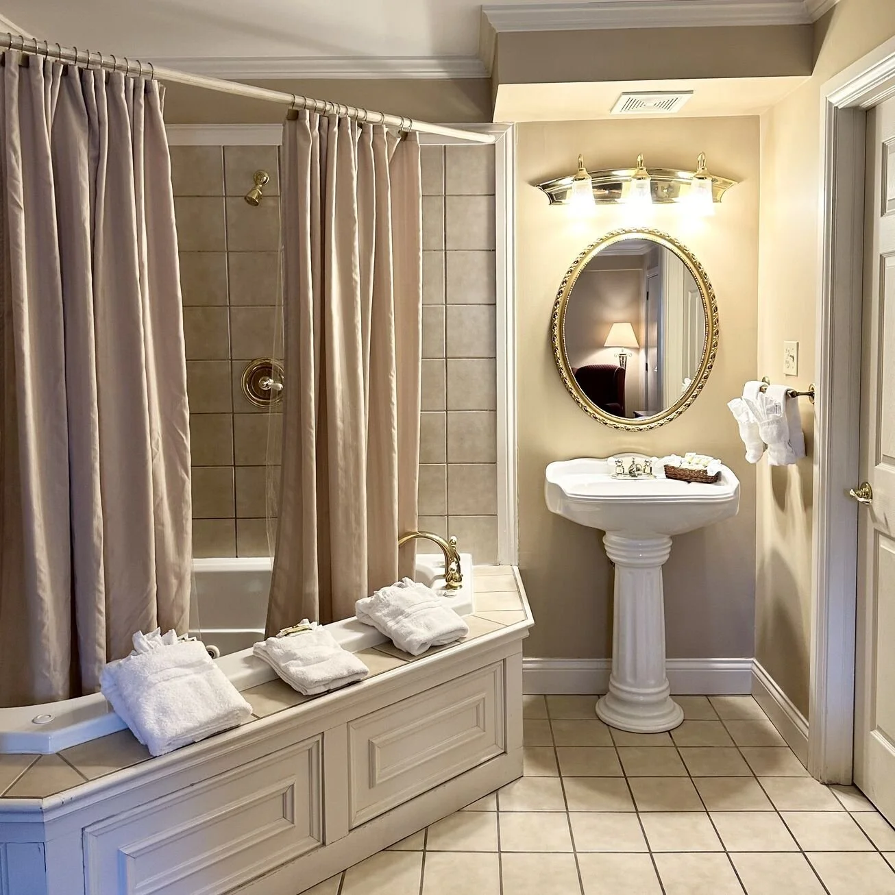 A bathroom with a bathtub enclosed by beige curtains, a pedestal sink with a mirror above it, and neatly folded white towels on the ledge and a towel rack.