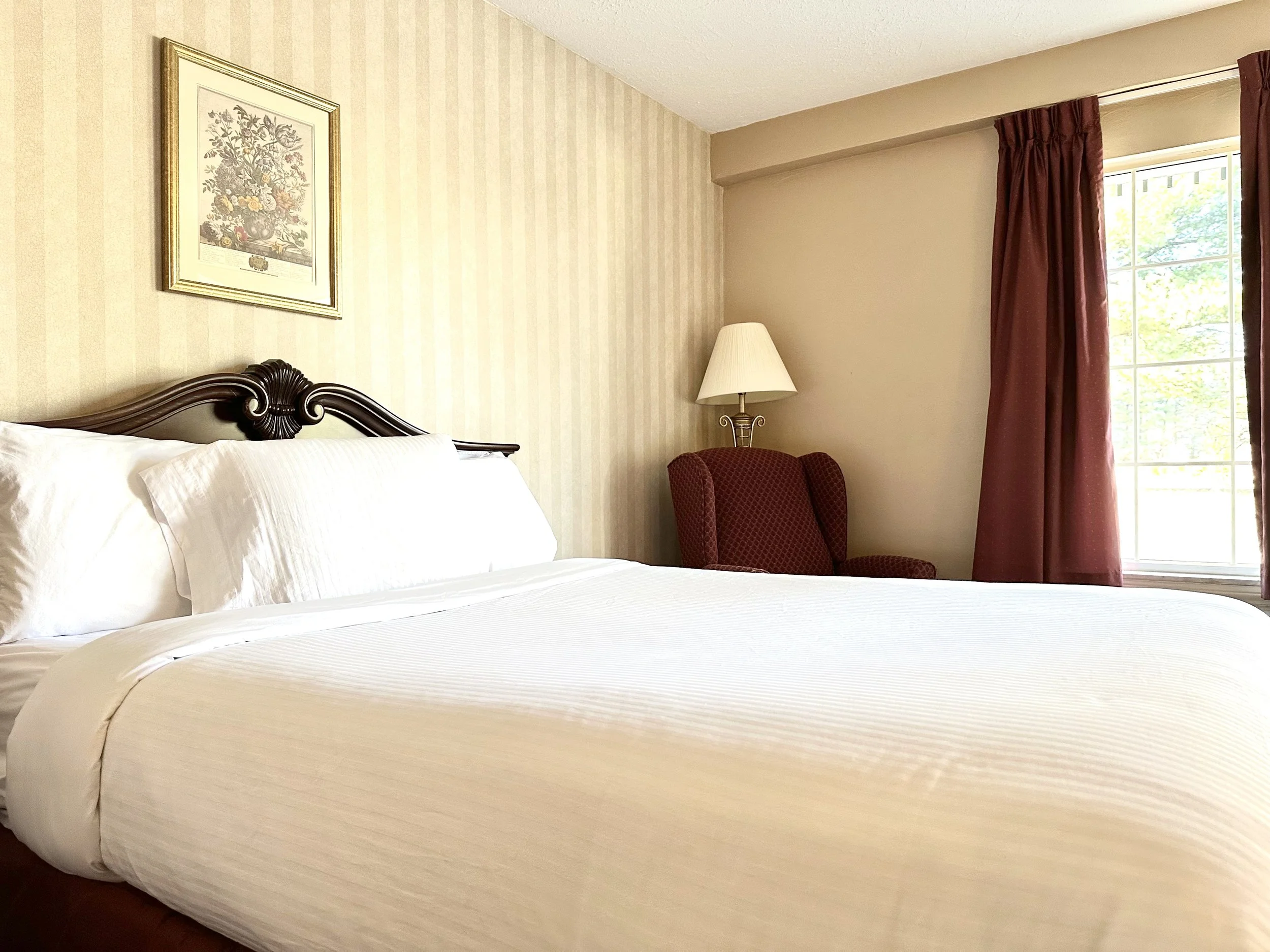 A hotel room with a large bed, white linens, and pillows, a side chair with a floor lamp, and a window with red curtains, beige walls, and artwork hanging above the bed.