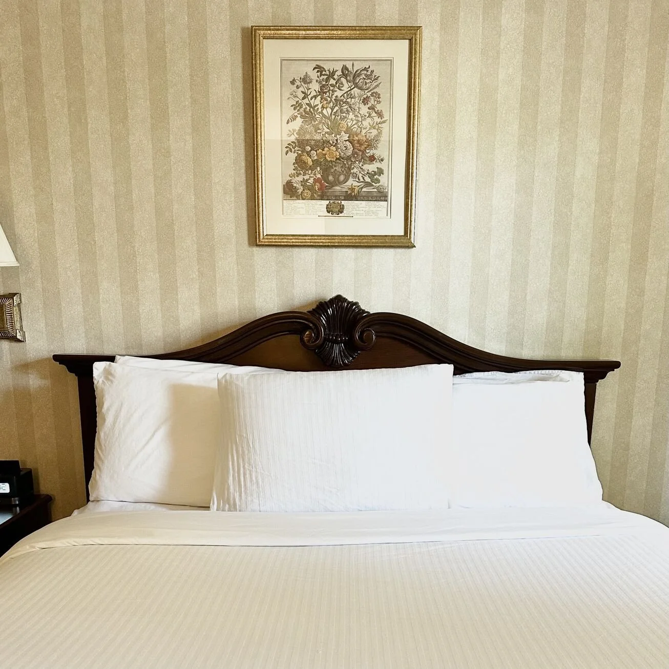 Neatly made hotel bed with white pillows and a cream-colored bedspread, wooden headboard, above framed floral artwork, beige striped wallpaper.