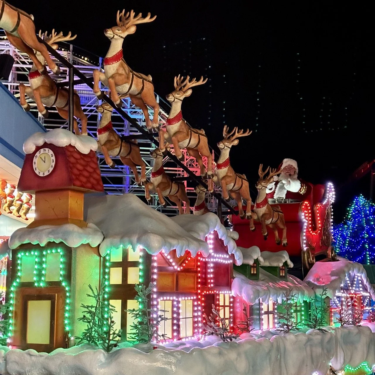 A festive ice skating rink decorated with Christmas lights at night, with a large illuminated Christmas tree at center, surrounded by trees with colorful lights, and a tall Christmas star at the top of a structure resembling a Christmas tree. A sign reads "Kings Island Winterfest."