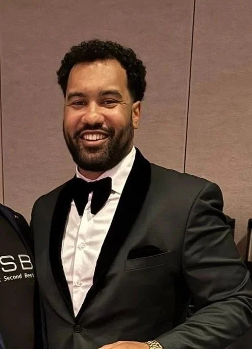 Man with curly dark hair and beard, dressed in a black tuxedo with a white shirt and black bow tie, smiling at the camera indoors.