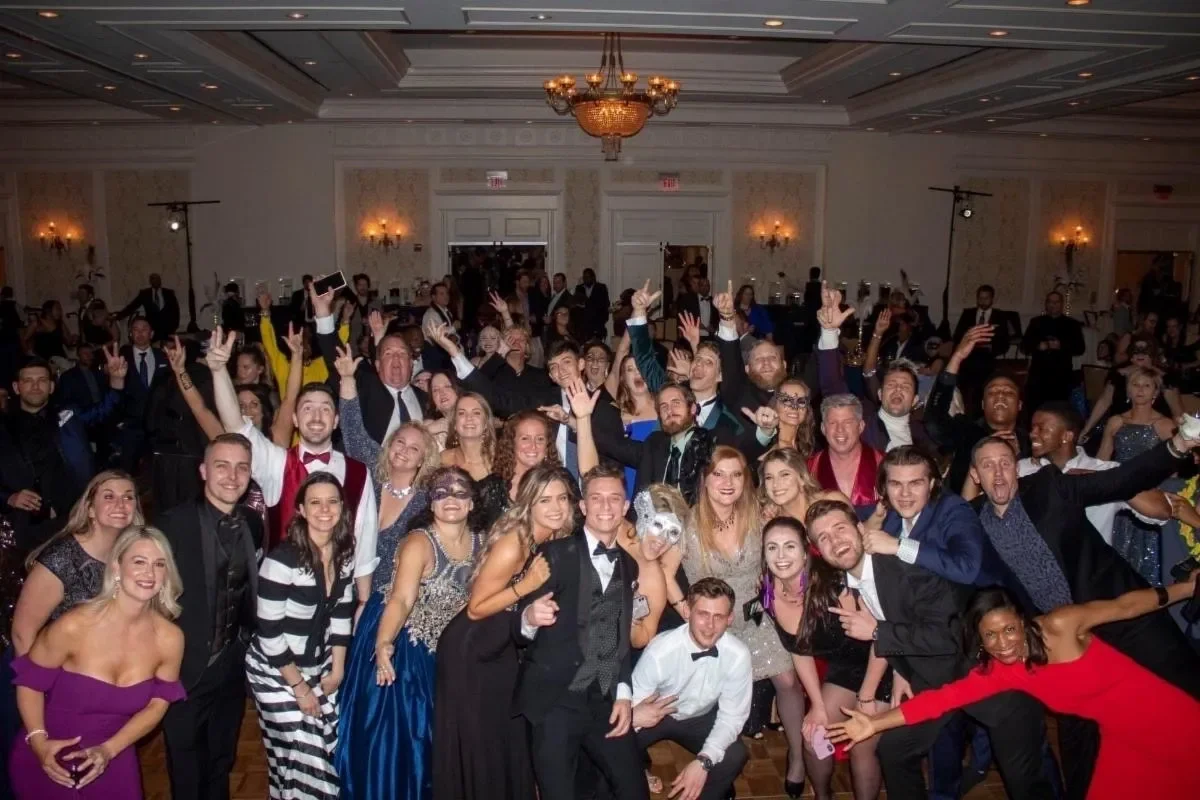 Group of people at a formal event or party in an elegant ballroom, smiling and celebrating, some wearing masks and costumes.