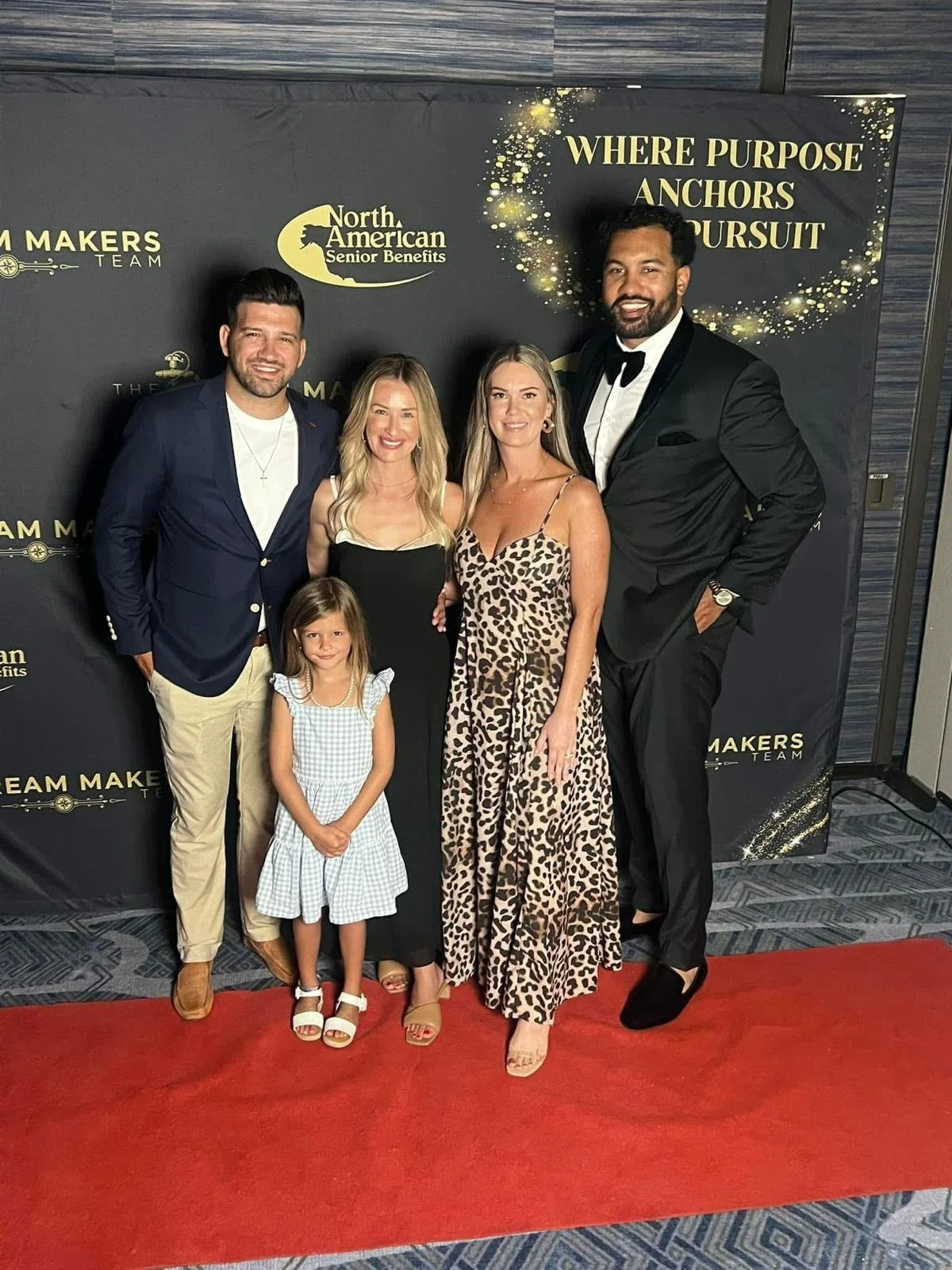 Group of five adults and one child standing on a red carpet at an event with a black backdrop that reads 'WHERE PURPOSE ANCHORS PURSUIT' and 'North American Senior Benefits'. The people are dressed in semi-formal to formal attire, smiling at the camera.