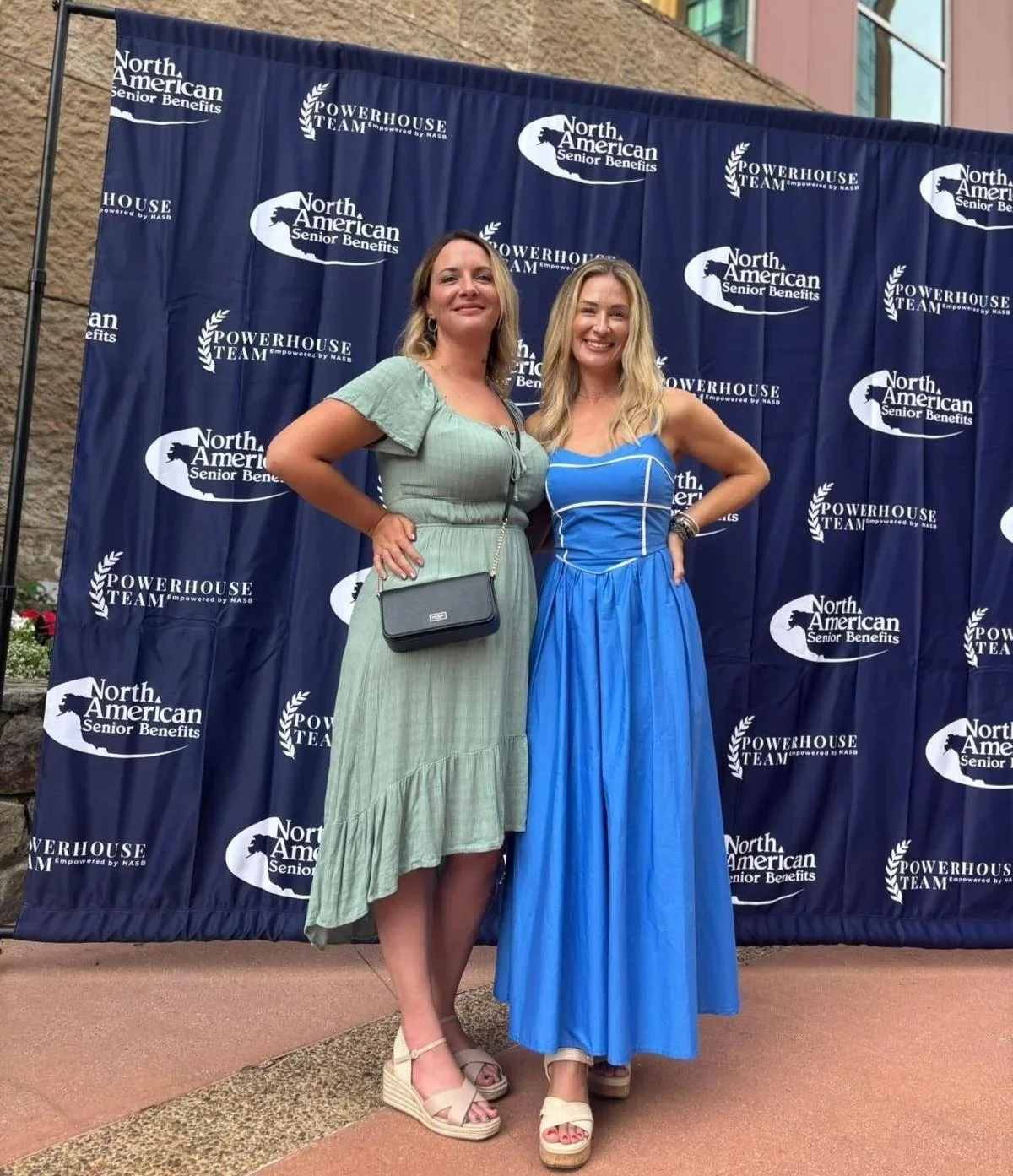 Two women pose in front of a navy blue backdrop with white logos for North American Senior Benefits and Powerhouse Team, smiling and standing side by side at an outdoor event.