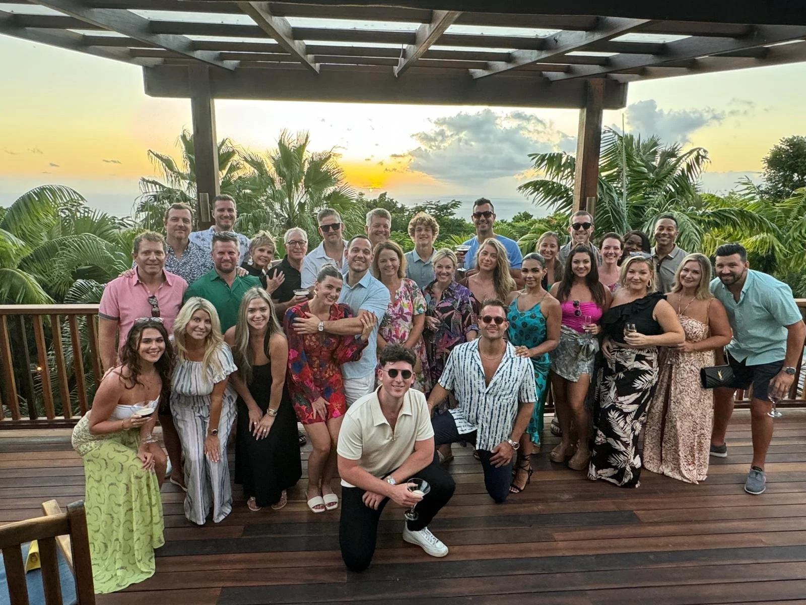 A large group of people at a sunset gathering on a wooden deck surrounded by lush tropical greenery, some holding glasses, smiling for the camera.