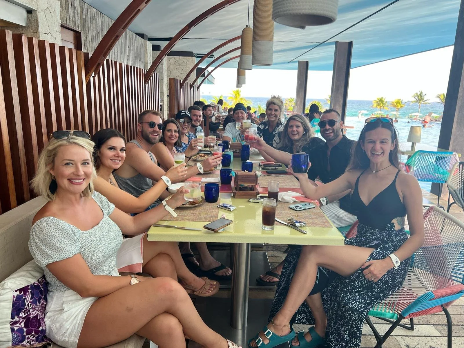 Group of people enjoying drinks at a seaside restaurant with an ocean view, palm trees, and umbrellas in the background.