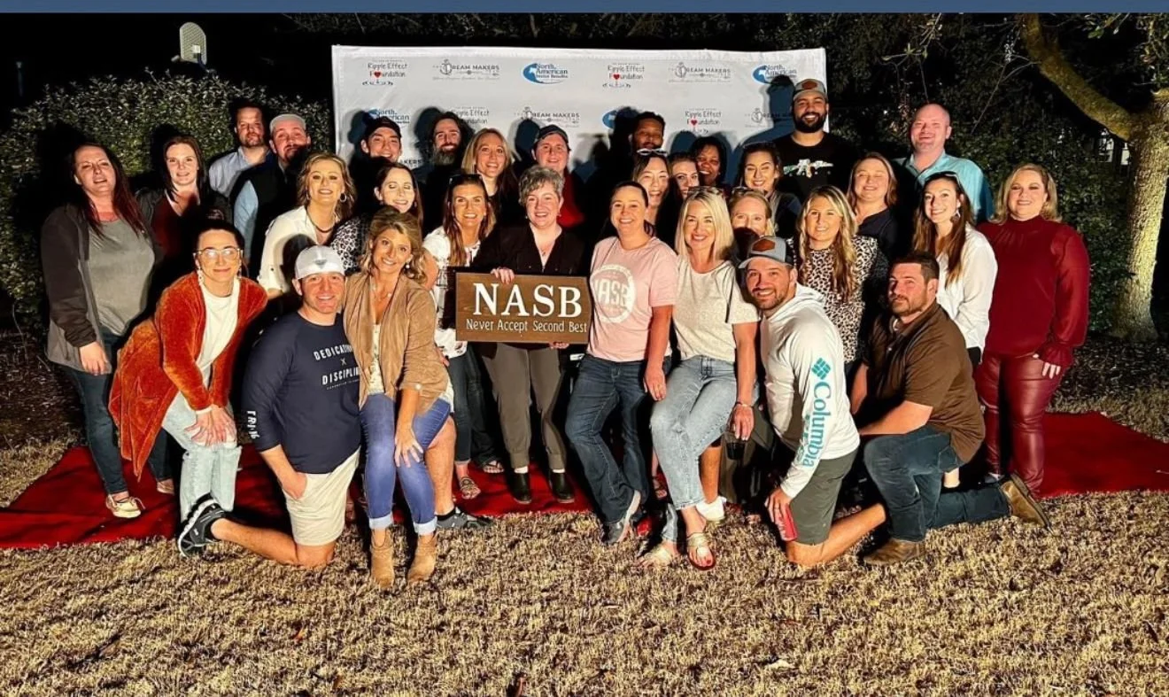 Group of people outdoors at night standing on a red carpet, with a step and repeat banner in the background, holding a NASB sign that reads "Never Accept Second Best."