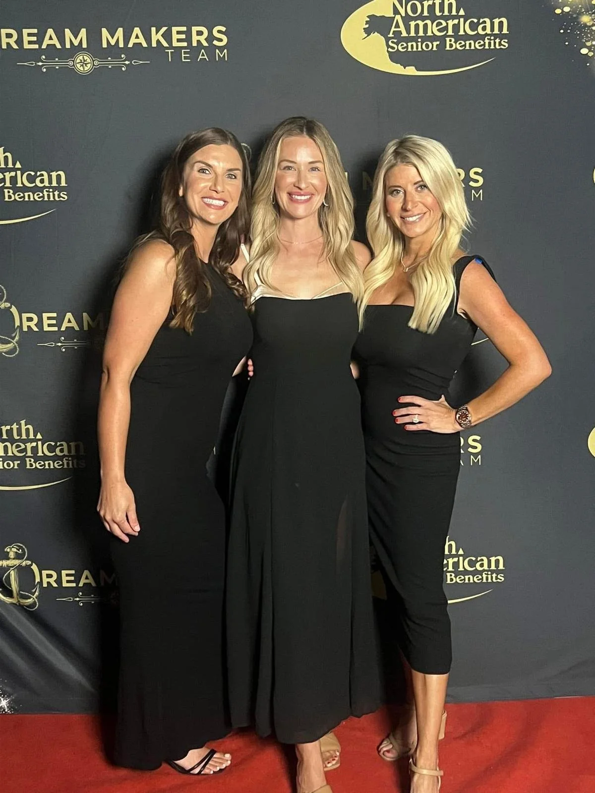 Three women in black dresses posing on a red carpet at an event with a backdrop displaying logos and text about North American senior benefits and Dream Makers Team.