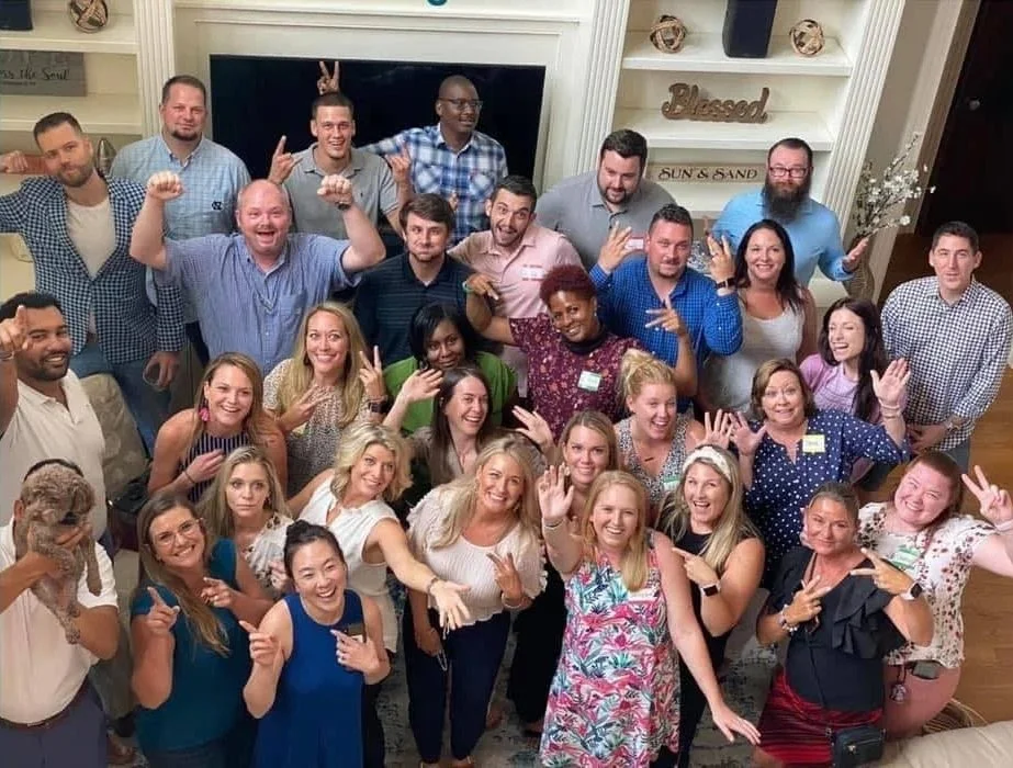 A large group of people gathered indoors, smiling and posing for a photo. Some are making gestures and waving, with a cozy, decorated room in the background.