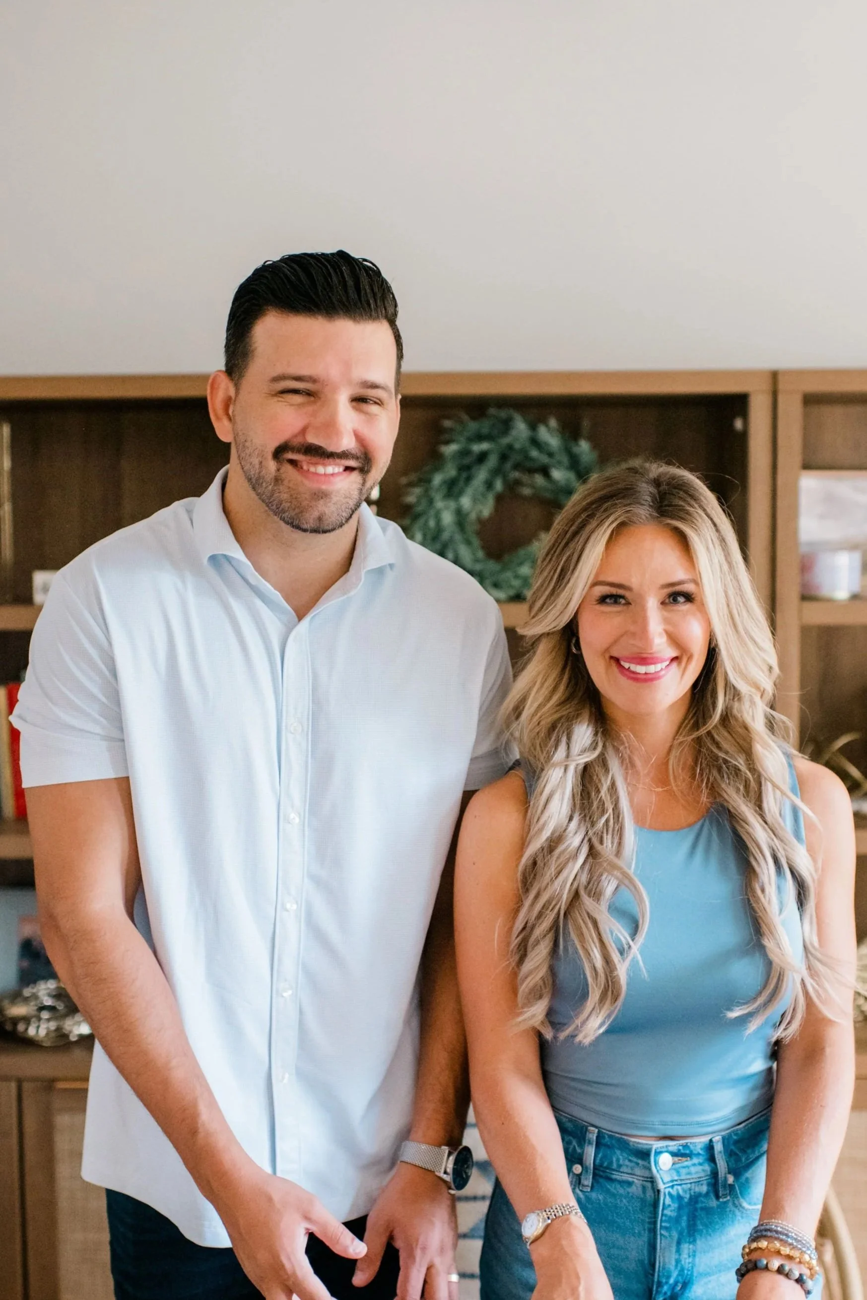 A man and woman standing side by side, smiling indoors with a holiday wreath in the background.