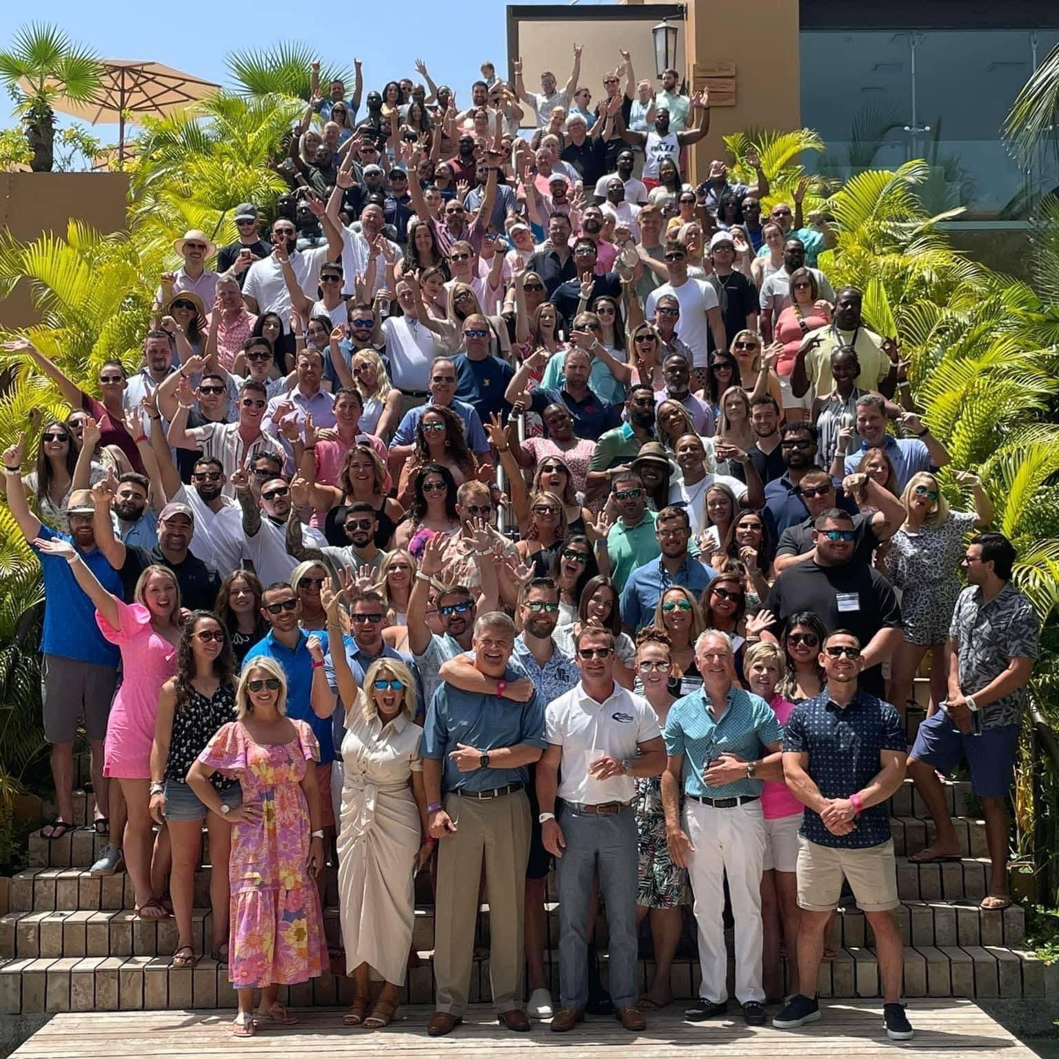 Large group of people gathered outdoors on stairs, smiling and waving at the camera, with palm trees and a building in the background on a sunny day.