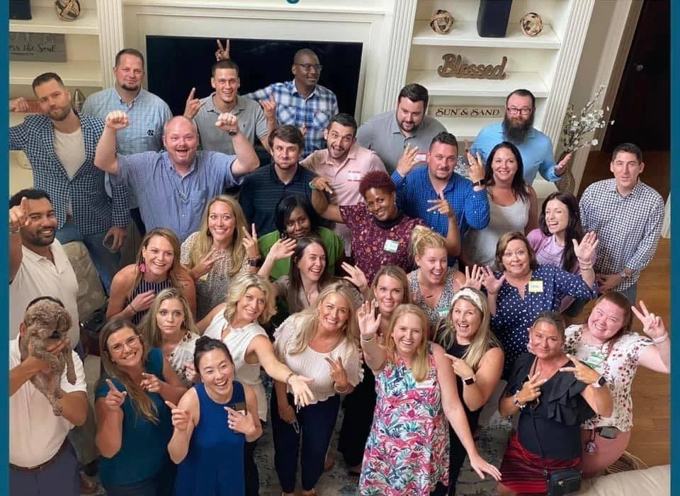A large group of people gathered indoors, posing for a group photo. They are smiling, making peace signs and celebrating.