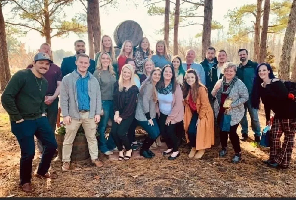 A large group of people posing outdoors in a wooded area during fall, standing around a large wooden wheel sculpture.