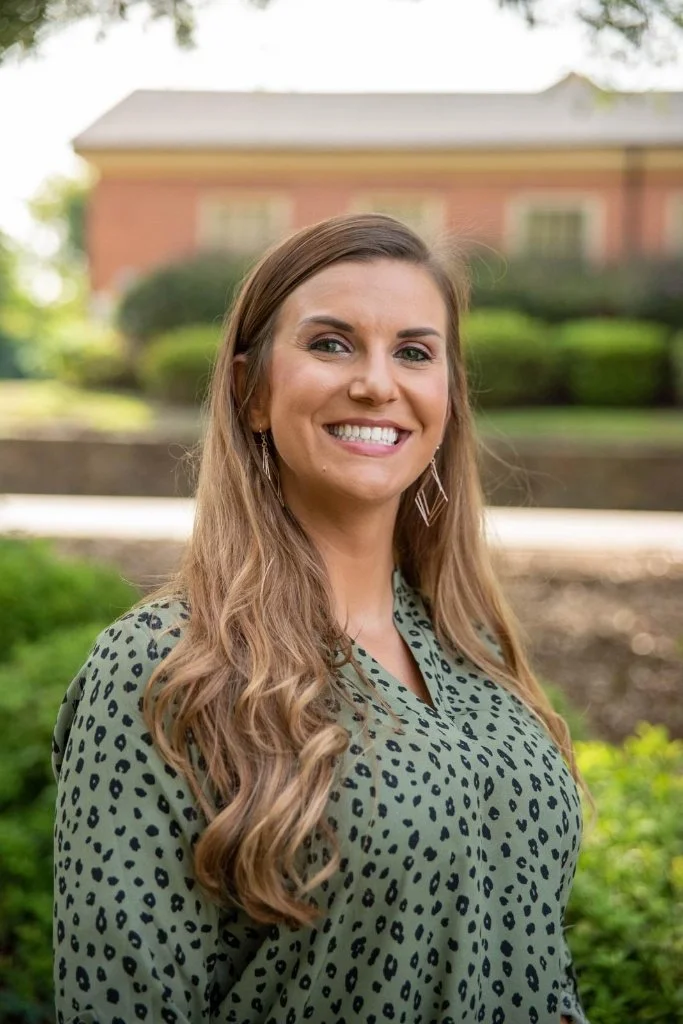 A woman with long wavy hair smiling outdoors in front of a red brick building and greenery.
