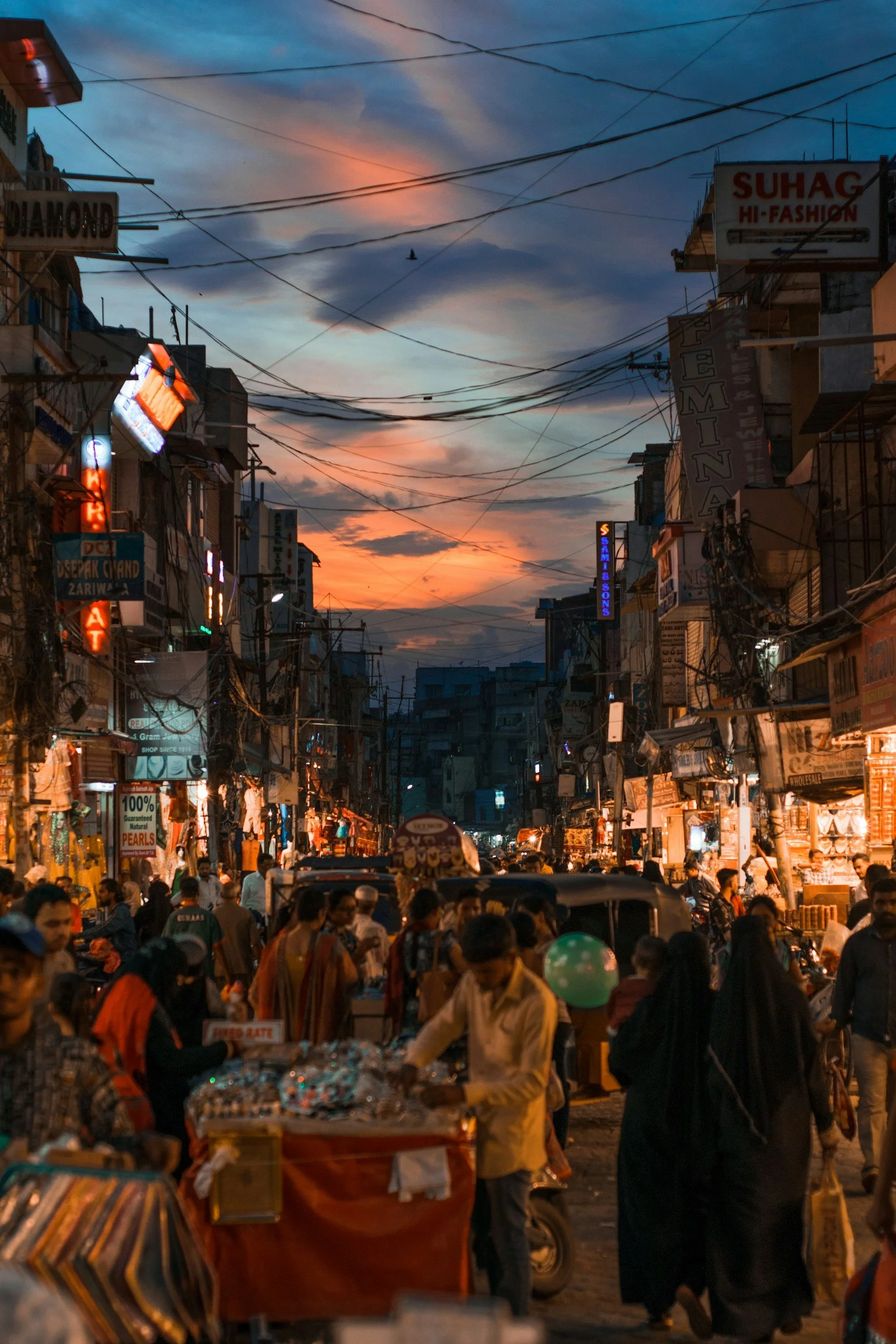 A busy street market at dusk with vendors and shoppers, illuminated signs, and a colorful sunset sky.