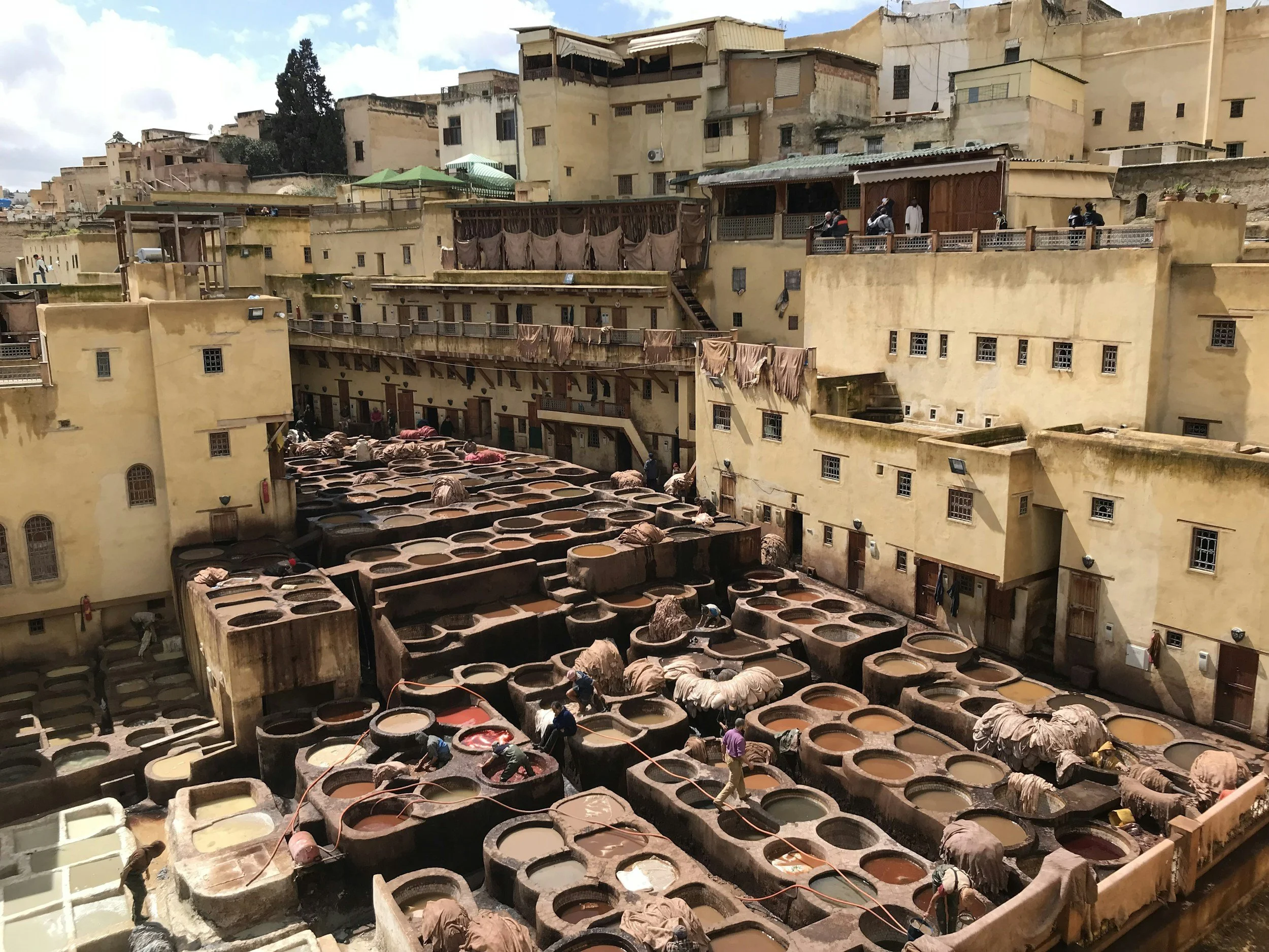 Traditional tannery with rows of large stone vats filled with various dyes and leather. Workers are seen processing leather in the open-air courtyard surrounded by old, yellowish buildings with balconies.