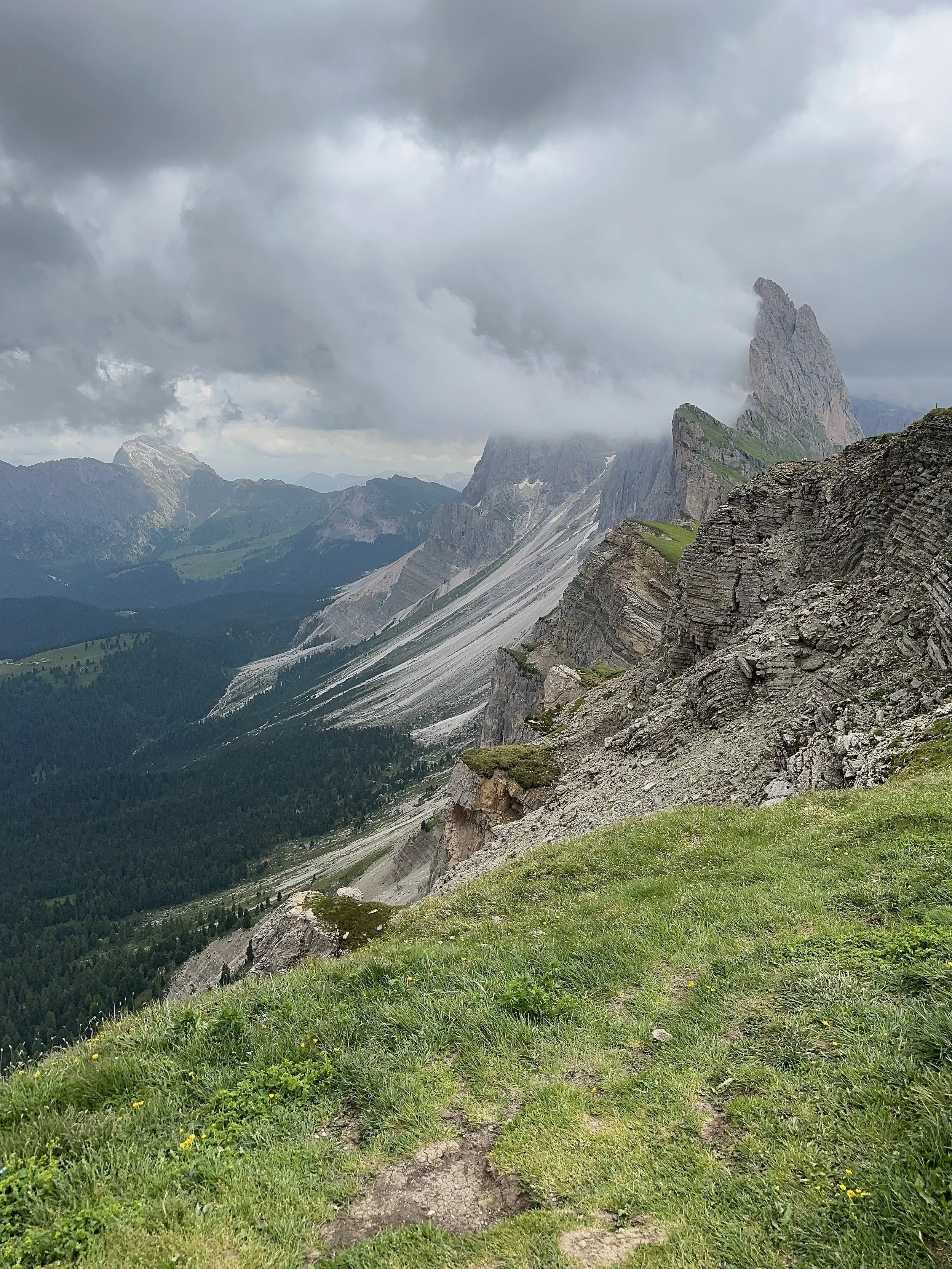 Mountain landscape with steep rocky cliffs, green grassy foreground, and cloudy sky.