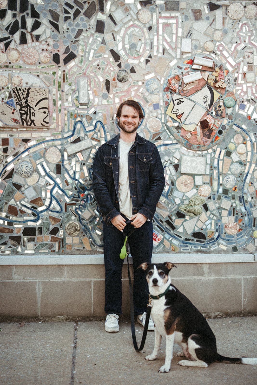 A smiling man with a beard and a dog sitting in front of a colorful mosaic mural featuring faces and abstract designs.