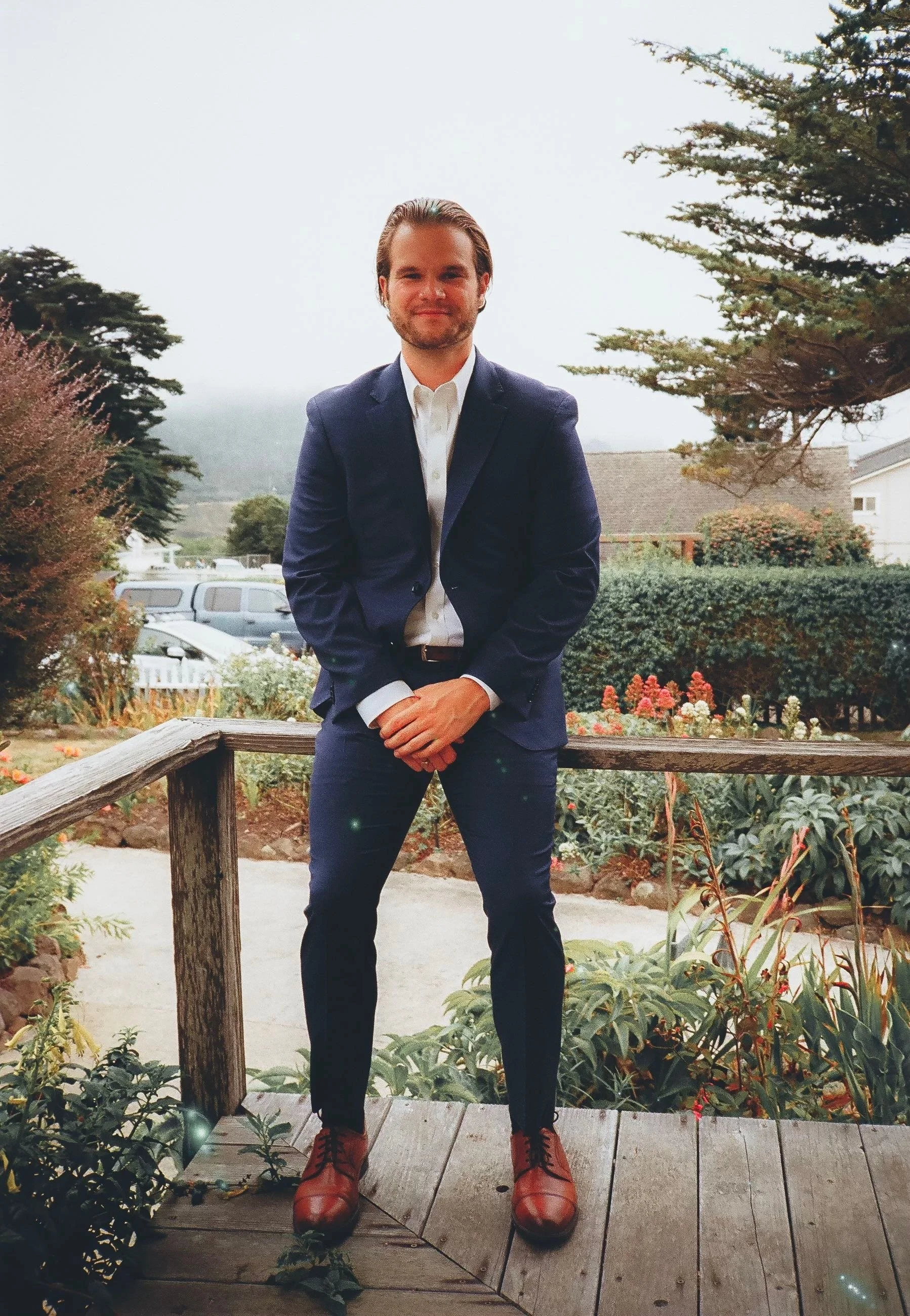 A man in a blue suit and brown shoes standing on a wooden porch with garden and trees in the background.