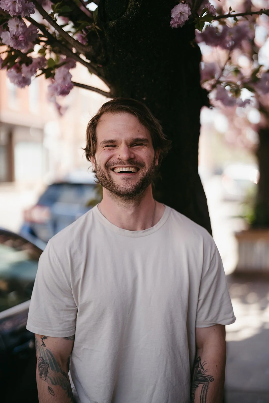 A smiling man with tattoos on his arms standing outdoors in front of a cherry blossom tree with pink flowers