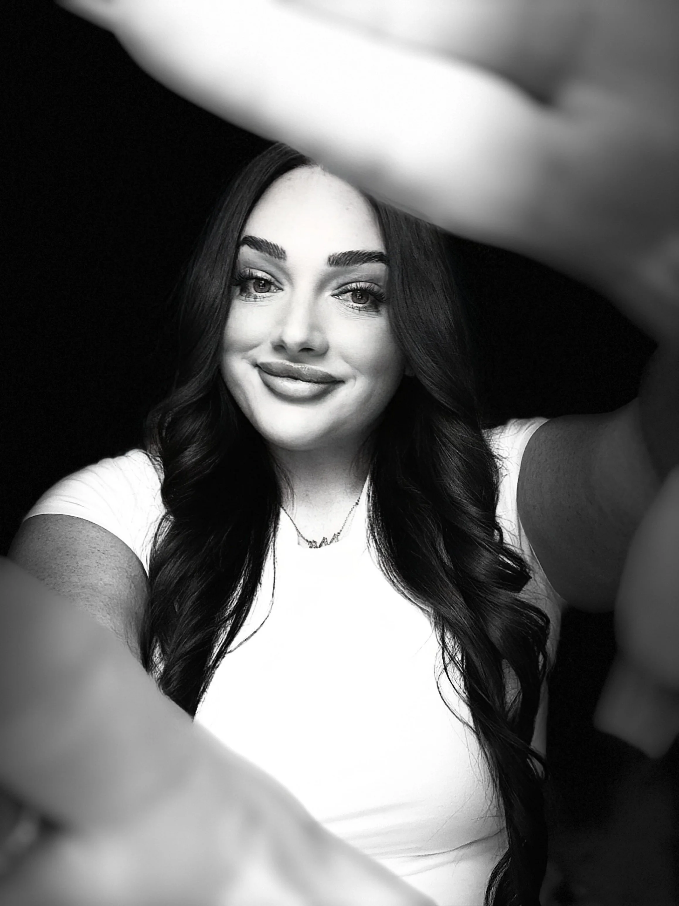 Black and white selfie of a woman with long dark wavy hair, smiling, wearing a white top and a necklace, with her hand reaching towards the camera.