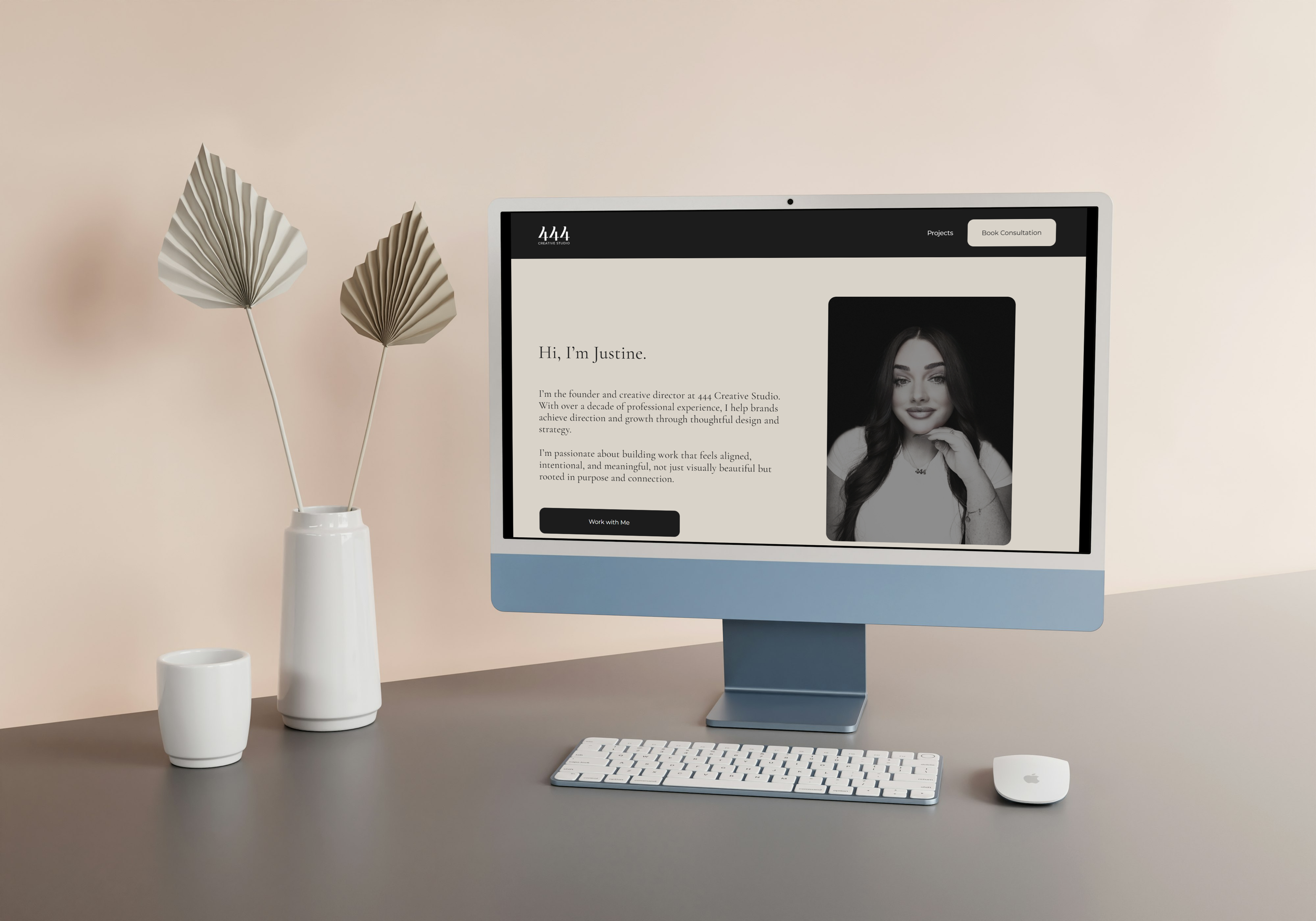 An iMac computer monitor displaying a website with a black and white photo of a woman and text introducing Justine, the founder of 444 Creative Studio, on a light-colored desk with a white keyboard, a white Magic Mouse, a white vase with decorative dried leaves, and a white mug.