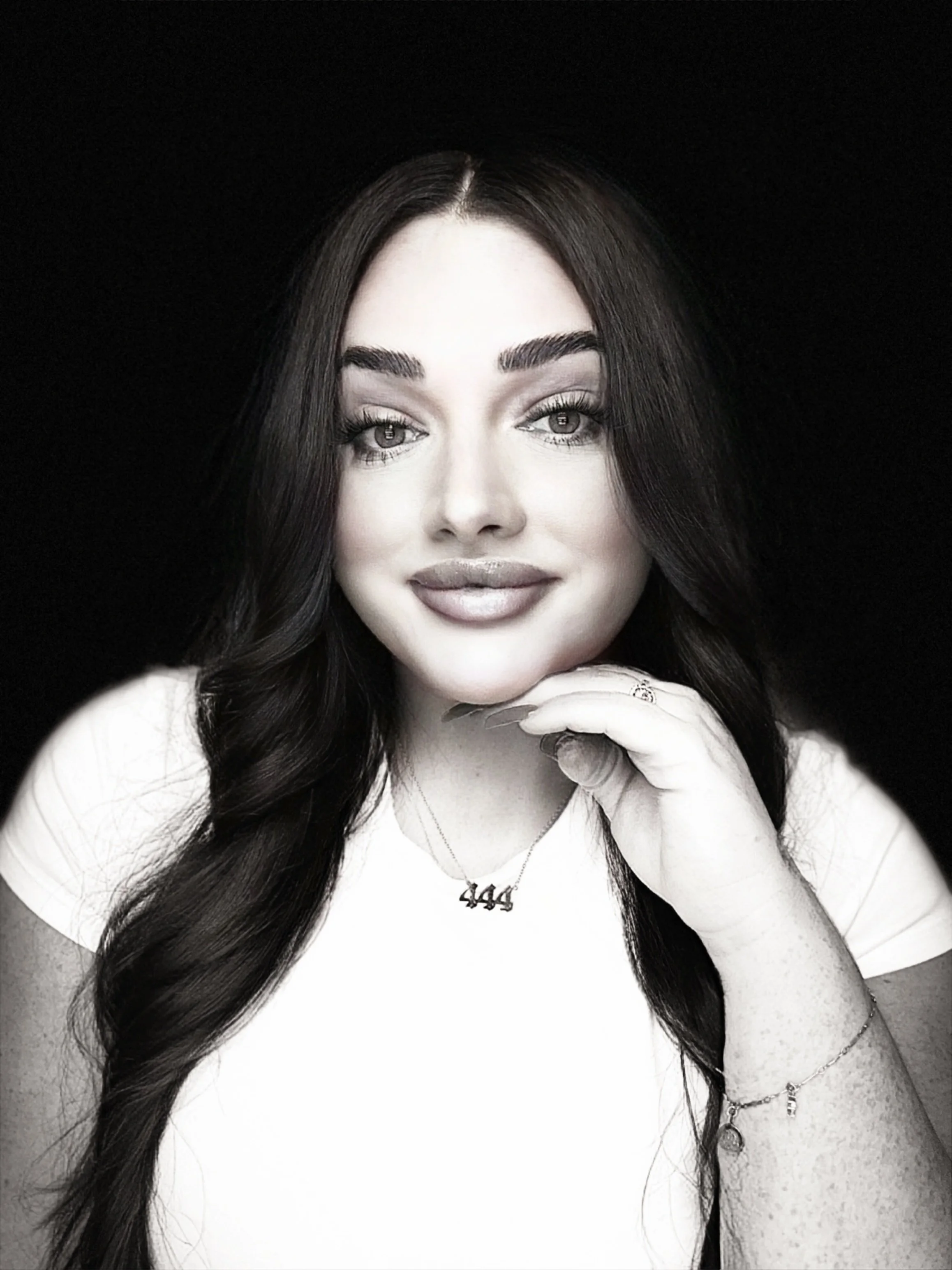 Black and white portrait of a woman with long dark hair, smiling, with her hand resting on her chin, wearing a white top and jewelry including a necklace and bracelets.