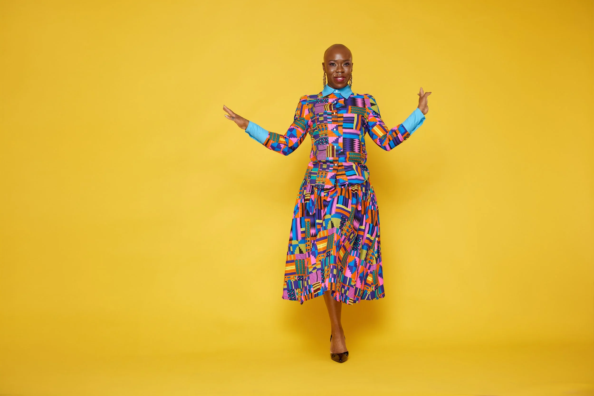 A woman stands in front of a yellow background wearing a vibrant, multicolored, patterned pantsuit with a blue collar and cuffs, and black heels.