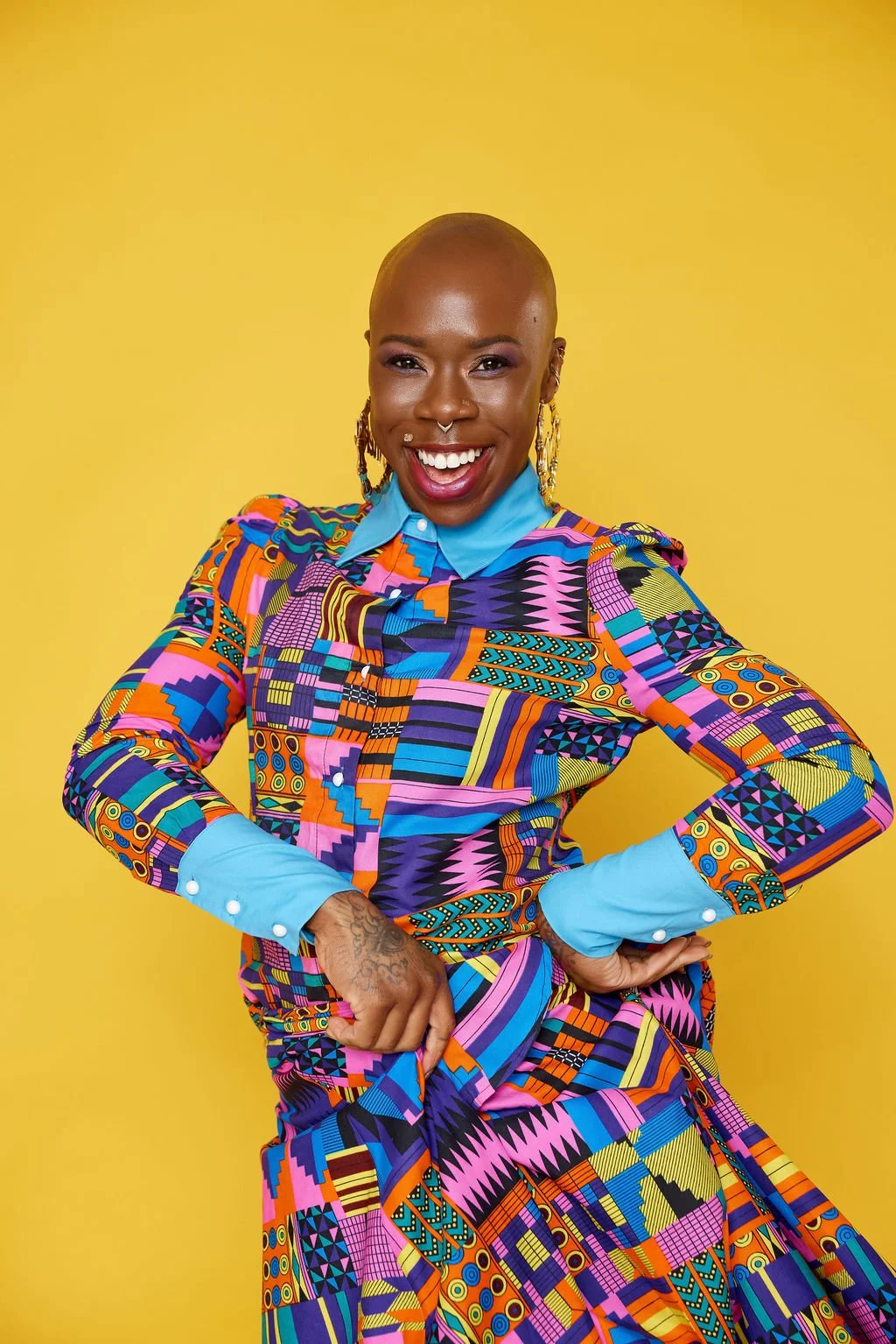 A smiling Black woman with a shaved head wearing a colorful, patterned dress with a light blue collar and cuffs, standing against a yellow background.