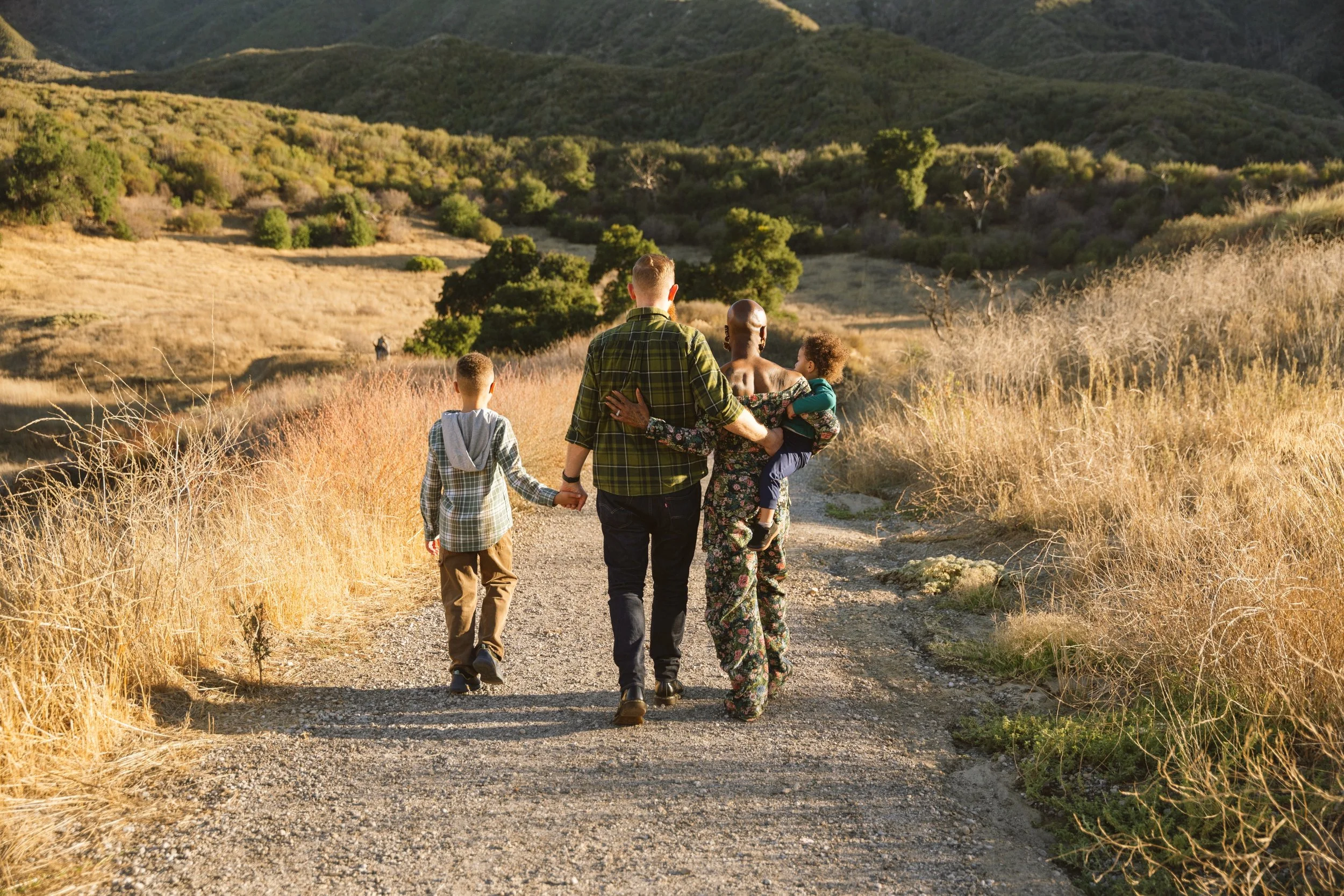 A diverse family of four walking on a gravel trail through a grassy, hilly landscape during sunset. The family includes two adults, one child holding hands with one adult, and another child being carried by one of the adults.
