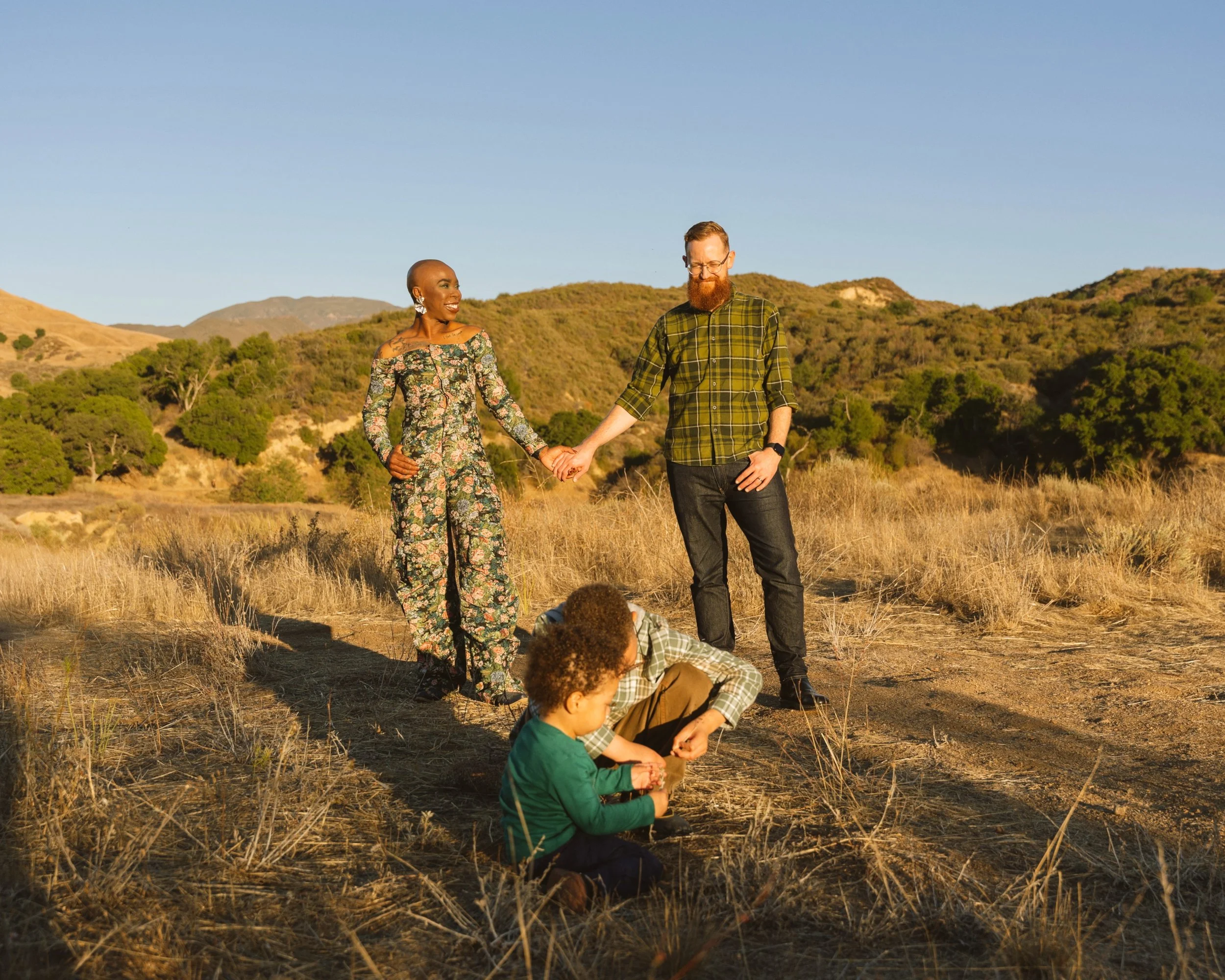 A diverse family of five holding hands and playing outdoors in a grassy field with hills in the background during sunset.