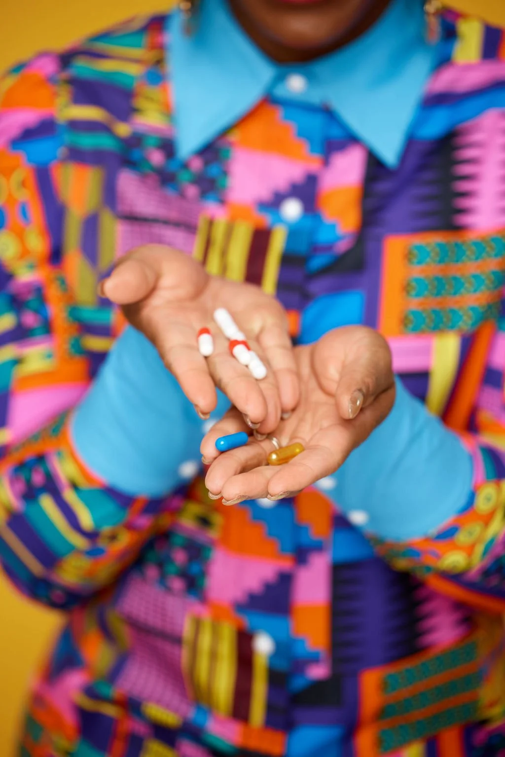 Person holding various colorful pills and capsules, dressed in a vibrant, patterned shirt with a blue collar.