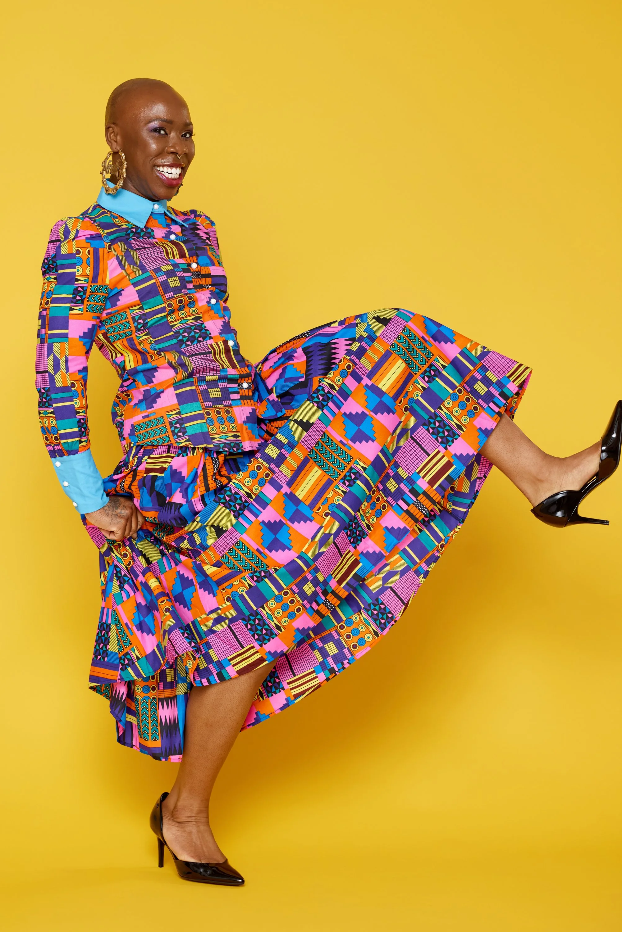 A woman with a shaved head smiling and lifting her leg, wearing a vibrant, colorful patterned outfit with black high heels, standing against a yellow background.