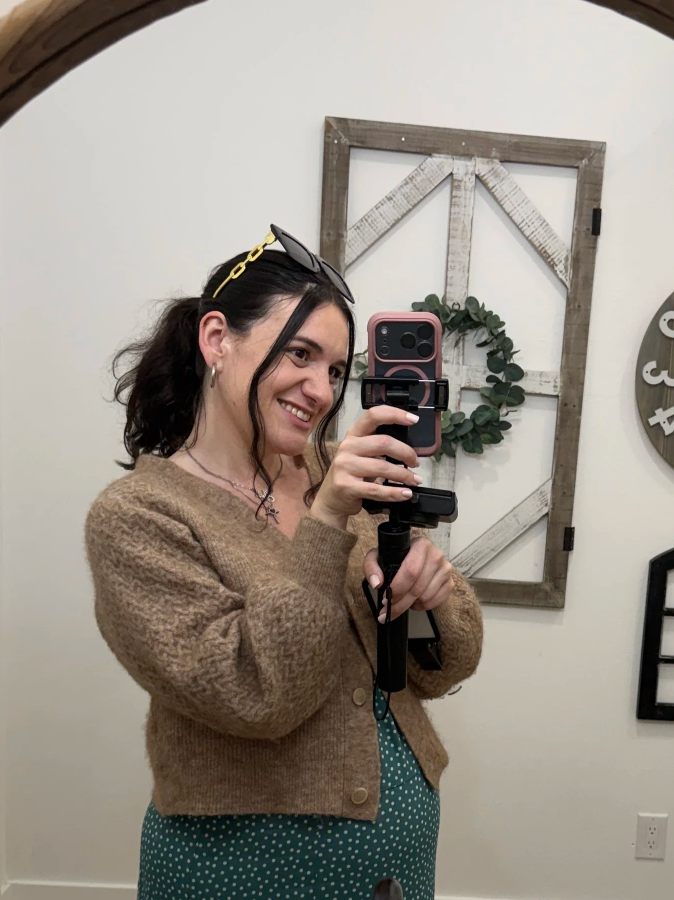 Woman taking a selfie in a mirror, wearing a brown cardigan and green dress with white polka dots, with sunglasses on her head and earrings.