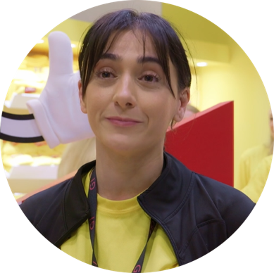 A woman with dark hair and bangs smiling, standing indoors with a yellow background. She is wearing a black jacket over a yellow shirt and has a lanyard around her neck. There is a large, white glove prop and a red object behind her.