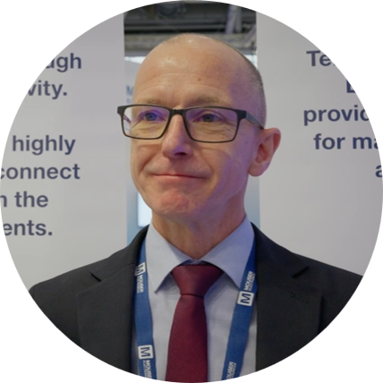 A man with glasses, wearing a dark suit, light blue shirt, and maroon tie, standing in front of a presentation display with text, at a professional event.