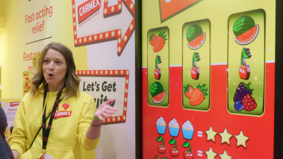 A woman in a yellow jacket stands next to a colorful game display featuring watermelons, strawberries, and grapes, with a prize section showing cupcakes and stars.