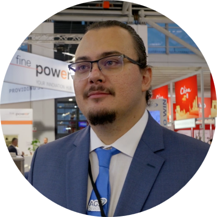 A man wearing a blue suit, white shirt, and blue tie with glasses, standing in an indoor exhibition or conference center.