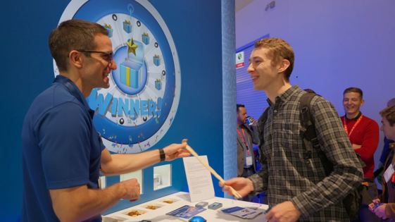 Two young men at a desk exchanging a wooden stick at an event or convention, with a large clock graphic and the word 'WINNER!' on the wall behind them, and other people in the background.