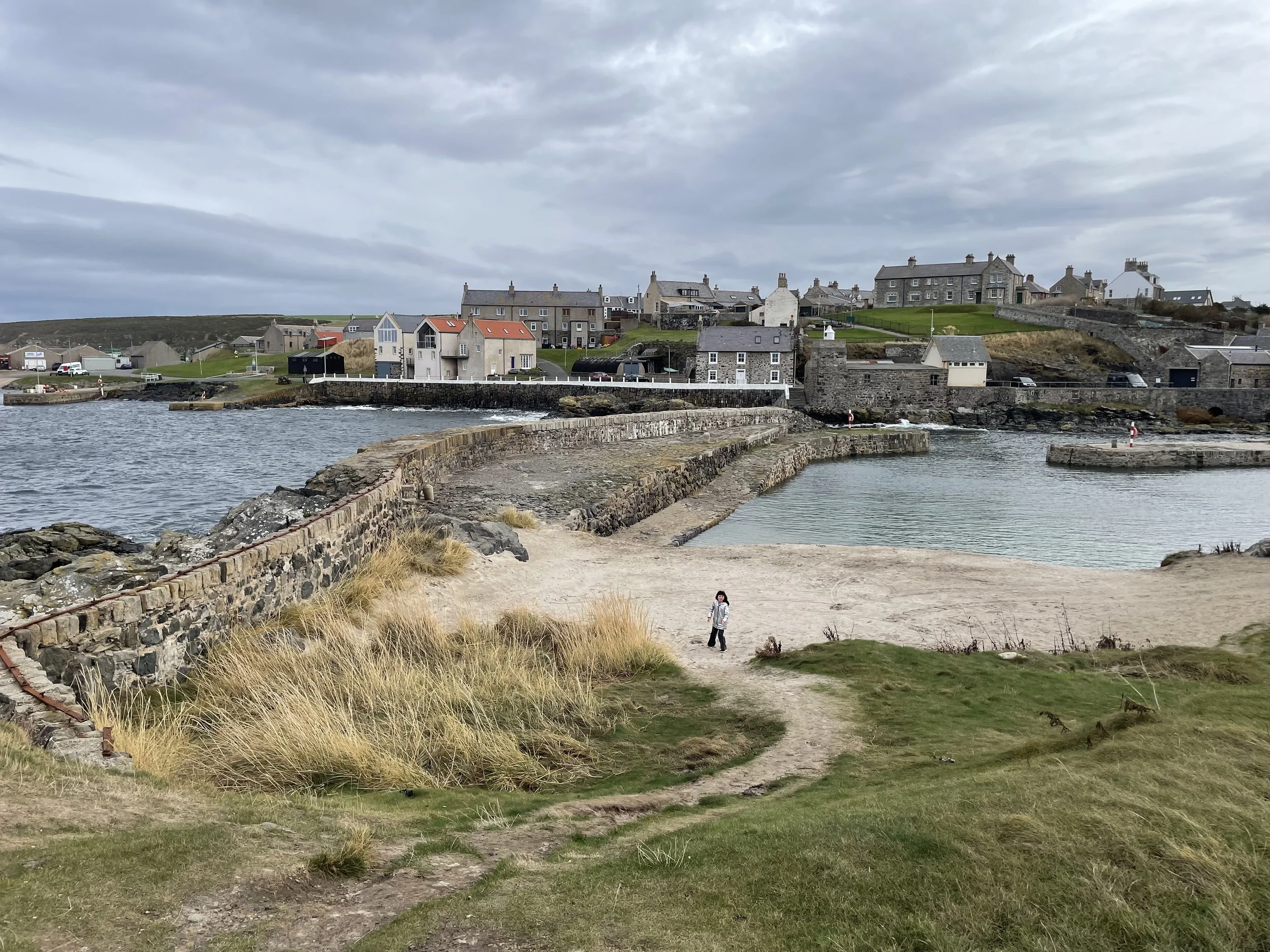 A child standing on a sandy path near the coast with grass and dunes in the foreground, stone walls, a harbour, and houses on a hill in the background under overcast skies.