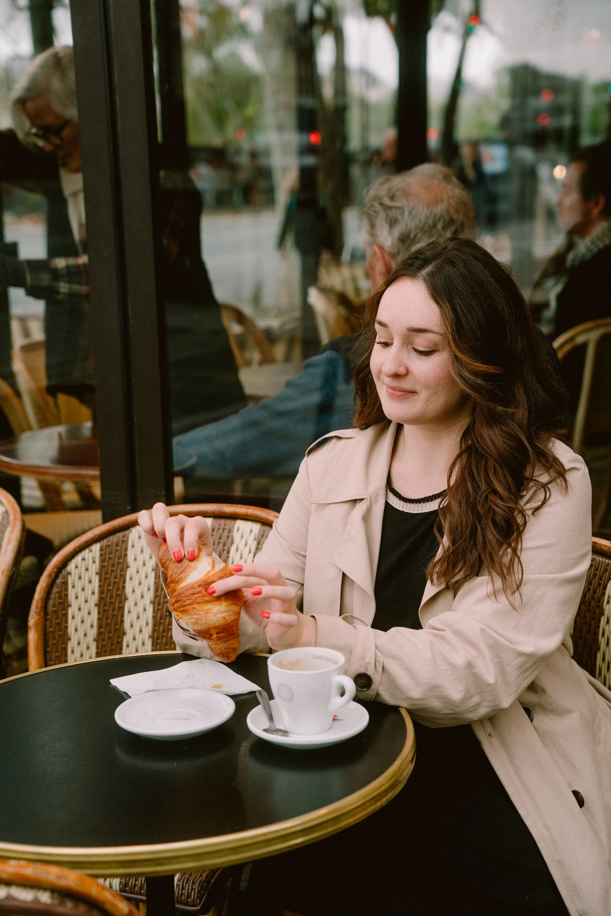 A woman with long wavy brown hair and a beige trench coat sitting at a table in a café, holding a croissant. There is a cup of coffee and an empty plate on the table.