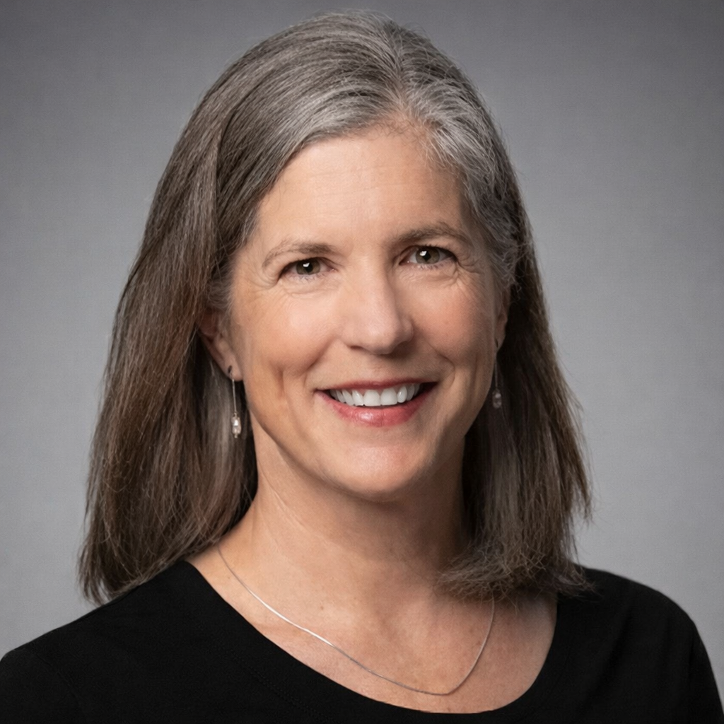 A middle-aged woman with long gray hair, smiling, wearing earrings and a black top, against a gray background.