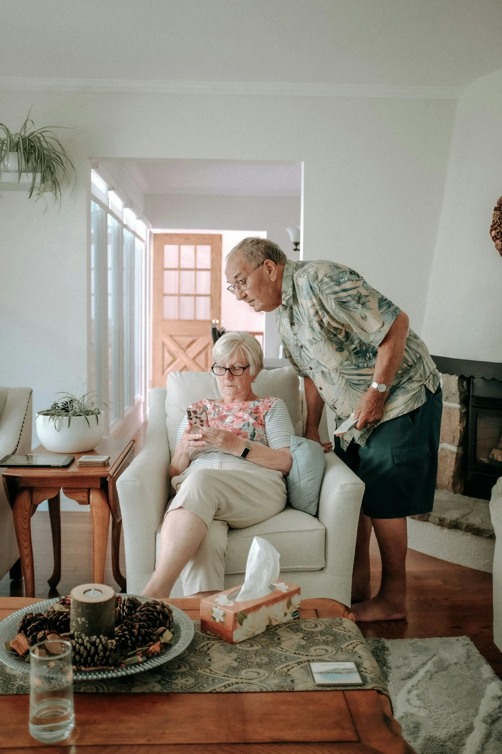 An elderly woman is sitting on a beige armchair, looking at her phone, while an elderly man leans over to see the phone. They are in a cozy living room with a fireplace, wooden door, and decorative items including tissue box, pinecones, and a candle.