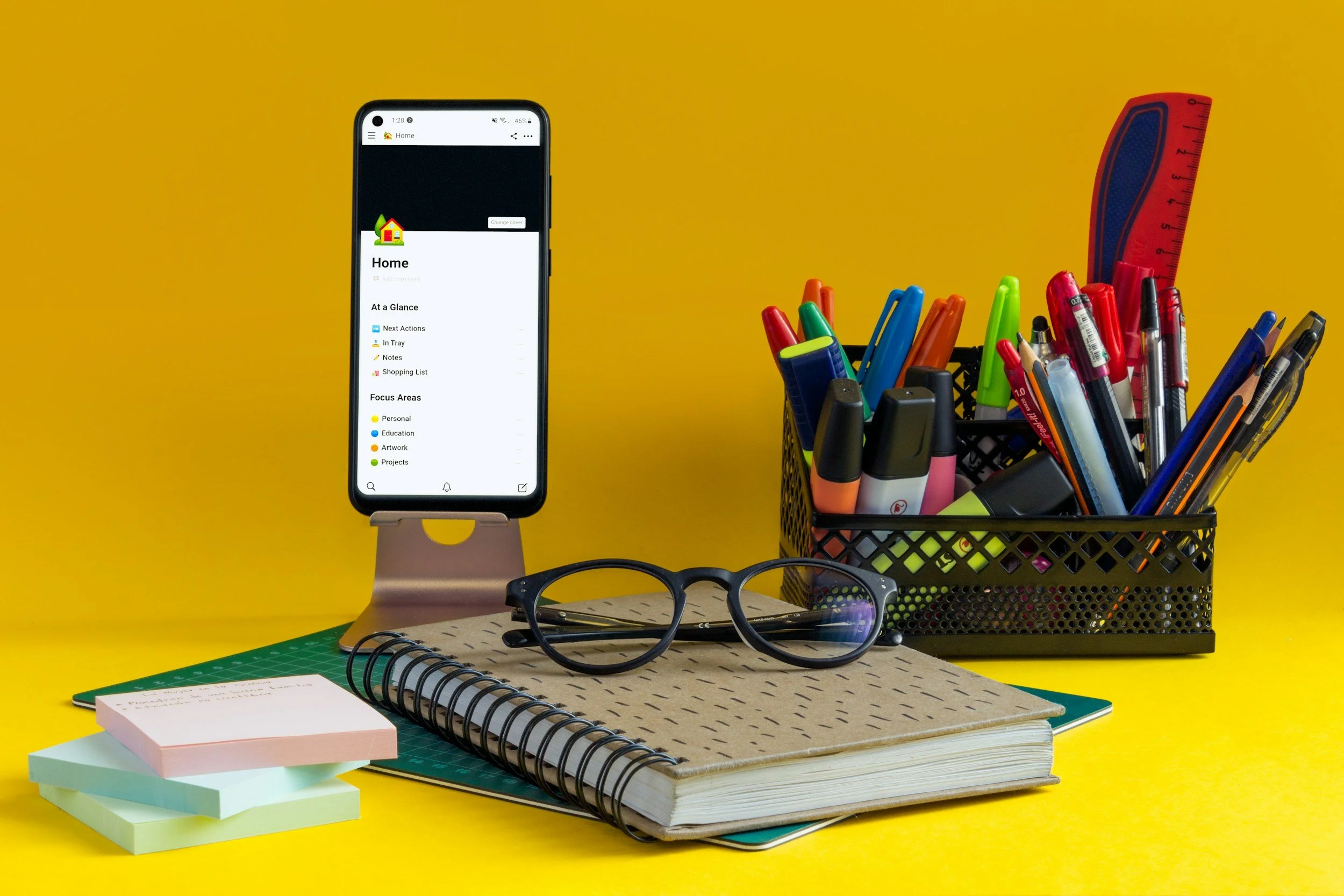 Desk with glasses, notebooks, sticky notes, a smartphone on a stand, and a black pen holder filled with colorful pens, markers, highlighters, and a ruler against a yellow background.