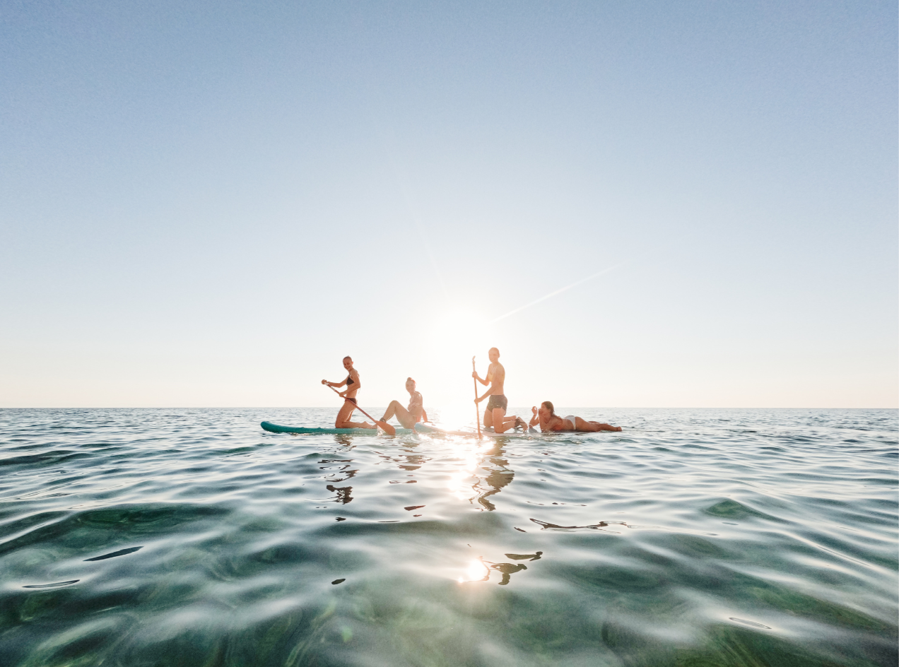 Four people enjoying a sunny day on the water, some on stand-up paddleboards and one lying on a surfboard, with the sun setting in the background.