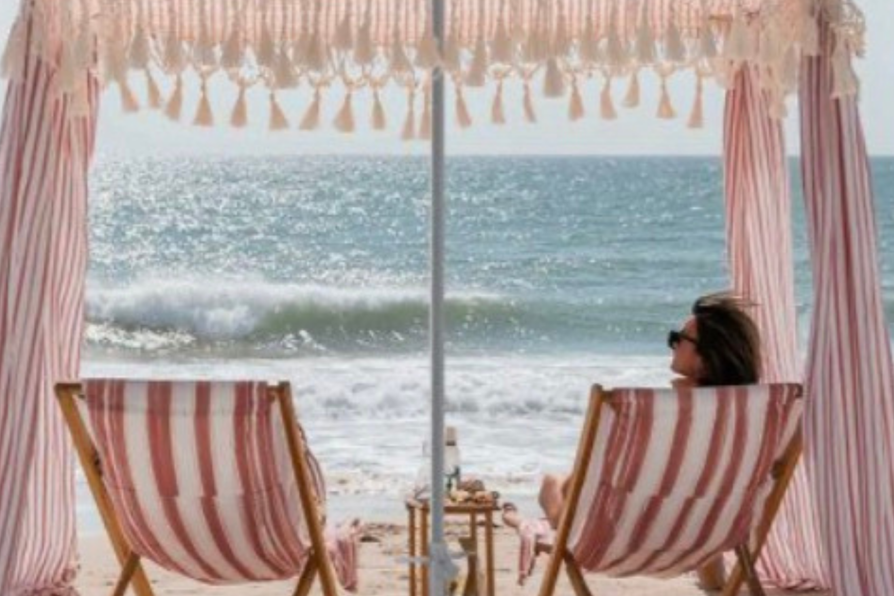 A woman sitting on a striped beach chair under a striped cabana, facing the ocean with waves, wearing sunglasses.