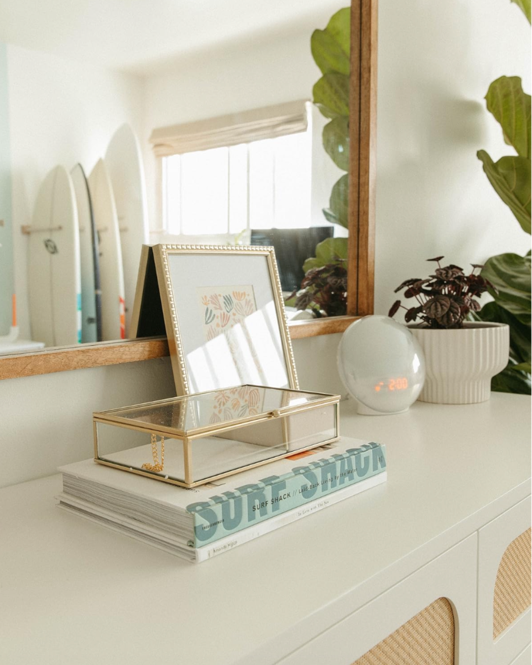 A white sideboard with decorative items including a framed artwork, a glass jewelry box, a stack of books about surfing, a white digital clock, and two potted plants, with a large mirror and window in the background.
