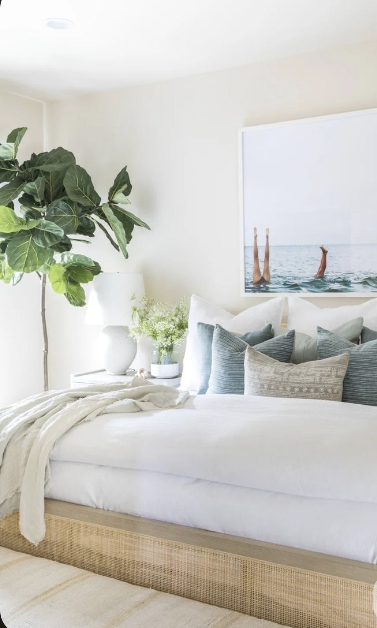 A bedroom with a bed featuring multiple pillows, a large houseplant, a white bedside table with a vase and flowers, and wall art showing two legs in the water.
